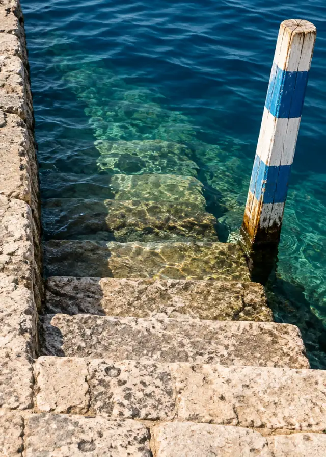 Stone harbour steps going down into clear blue water beside a striped tide pole.
