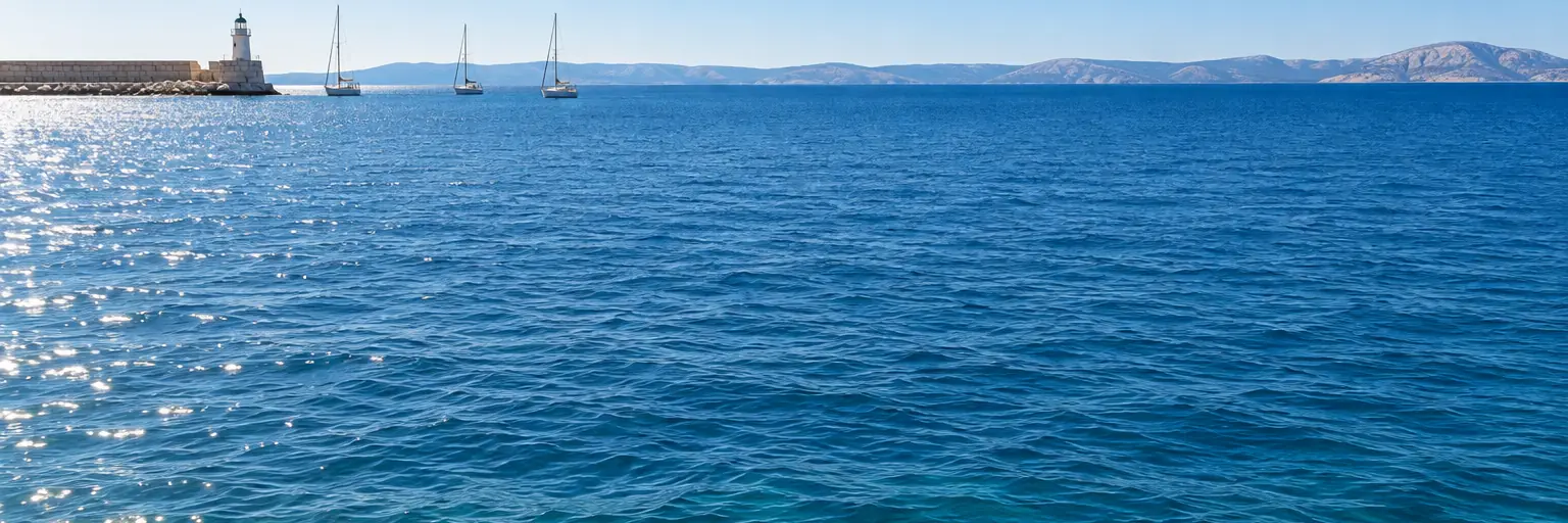 Calm blue harbour water with a small lighthouse and three sailing boats in the distance.
