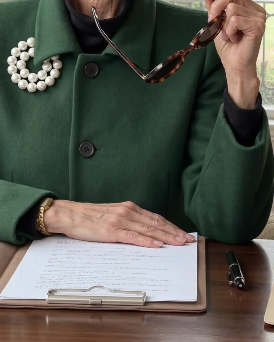 Maureen's hand resting on a page at her desk, a fountain pen beside it, her pearl brooch on the green coat above.