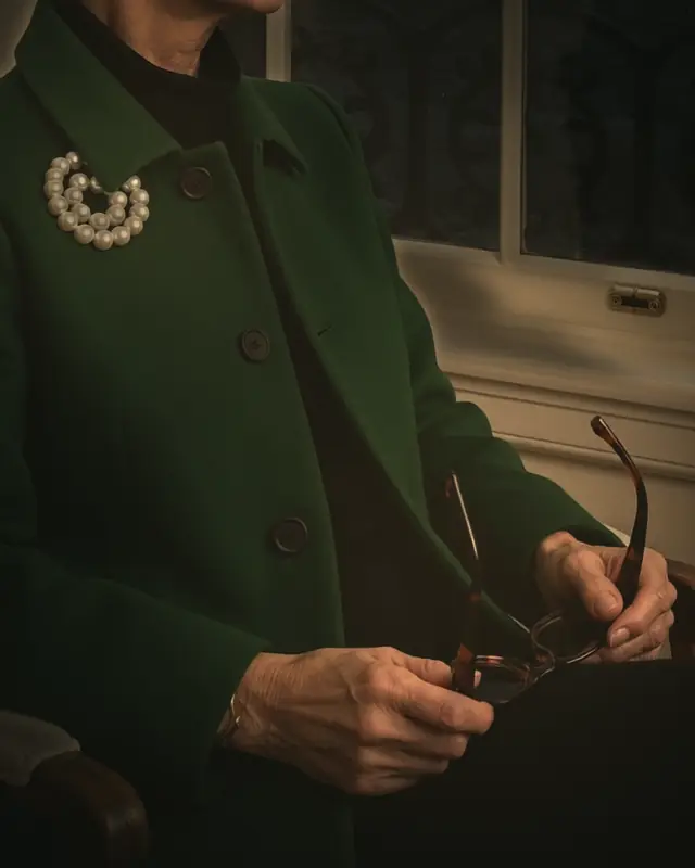 Maureen Pike's hands holding folded tortoiseshell sunglasses, in a money-green coat with a pearl brooch, by a window at night.